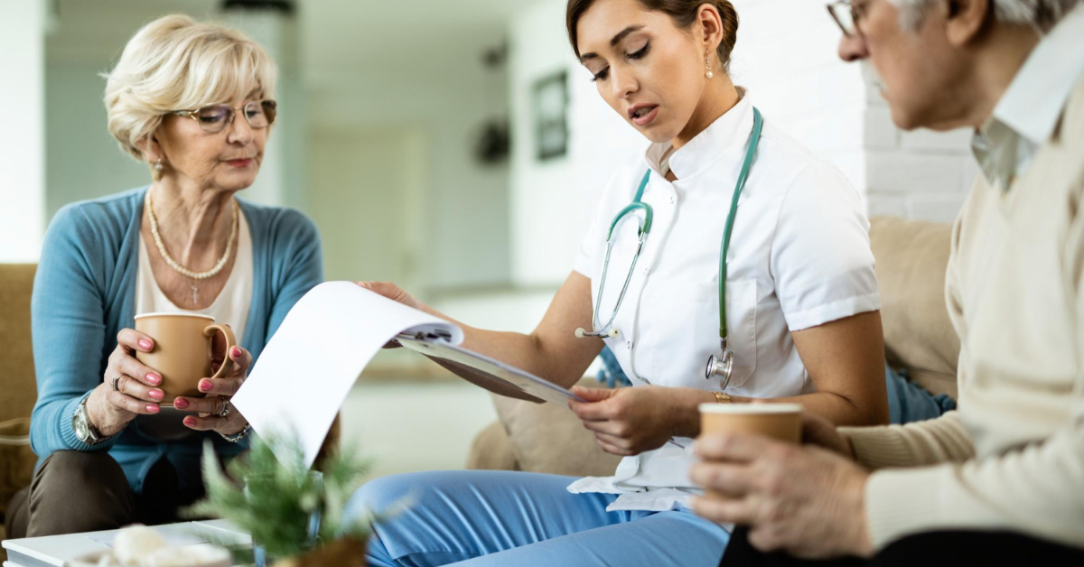 A healthcare professional discusses a document about elderly health insurance with two elderly patients, one man and one woman, as they hold coffee cups.