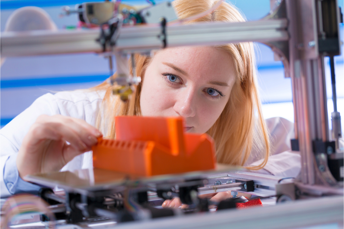 A person with blonde hair inspects an orange object being 3D printed within a machine, closely focusing on the innovative details of this advanced manufacturing process.