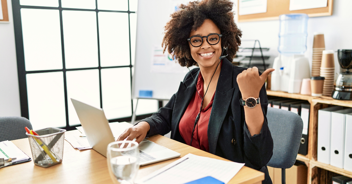 Woman with curly hair and glasses smiles while sitting at a desk. She is wearing a red top and black blazer, working on a laptop, and showing a thumbs-up. Office supplies and water are on the desk, helping tackle back office support challenges for small businesses.