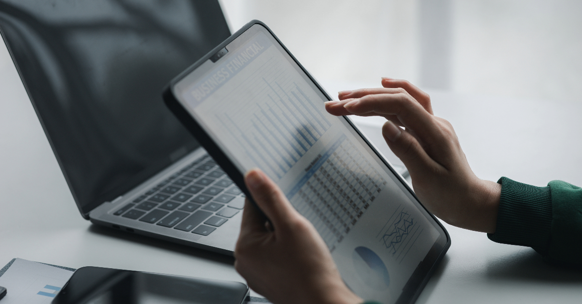 A person holding a tablet displaying business financial charts next to an open laptop on a desk, ensuring compliance with regulations.