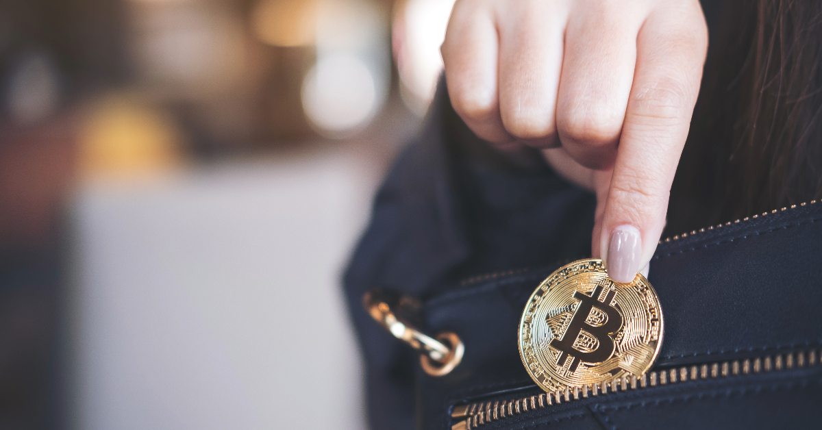 A hand is placing a gold Bitcoin coin into the zipper pocket of a black bag, highlighting the importance of secure storage in building a reliable crypto wallet infrastructure.