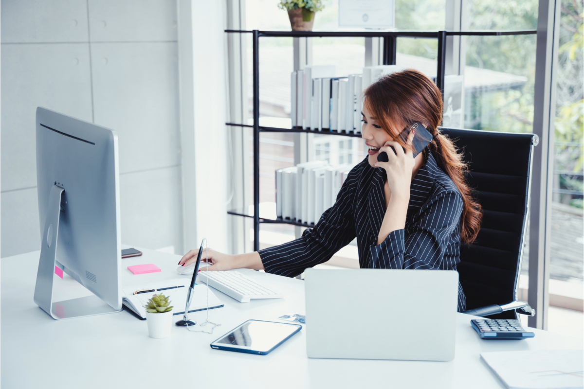 A person sitting at a desk in the digital age, cold calling on a phone while working on a computer. The desk hosts a desktop monitor, laptop, tablet, notebook, pen, calculator, and small potted plant. Bookshelves fill the background.
