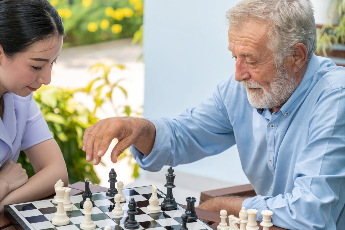 Two people, one elderly and one younger, are playing chess at an outdoor table. Demonstrating compassion and self-care, the elderly person moves a black piece while the younger person watches intently.