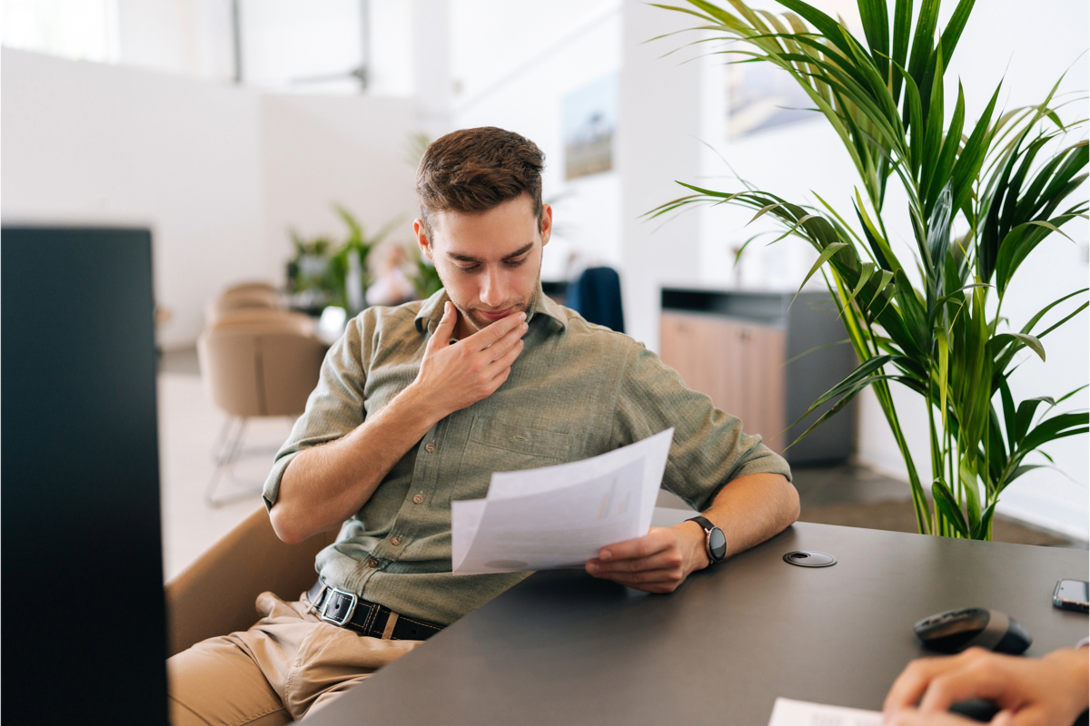 A man sits at a desk in an office, reading a document with a thoughtful expression. Beside him, there is a large green plant and part of another person is visible, potentially one of the accounting managers who help navigate the complex business landscape.