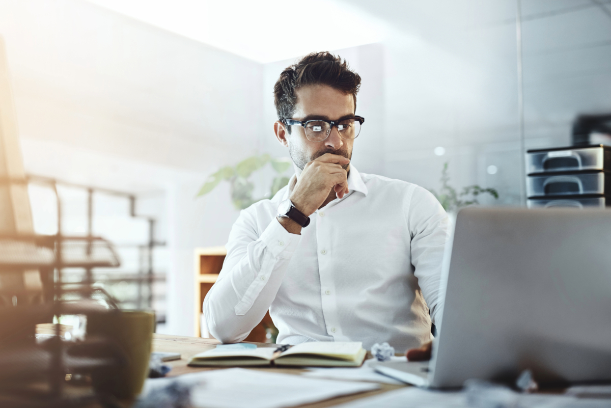 A man wearing glasses and a white shirt is sitting at a desk, looking at a laptop screen with a thoughtful expression. There are papers, a notebook, and a coffee mug on the desk as he considers navigating change amidst Google Algorithm Updates.