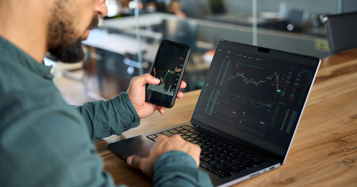 An investor sitting at a desk with a laptop and a cell phone, keeping track of Bitcoin prices in Australia.