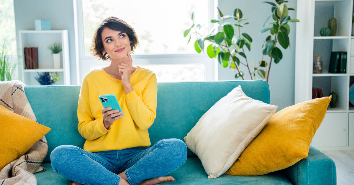 A person sits cross-legged on a turquoise sofa, holding a smartphone and looking thoughtful. The room is bright with plants and bookshelves in the background, perhaps drafting a step-by-step guide for their TikTok username or brainstorming crafting ideas.