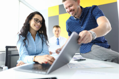 Three people in a bright office setting are engaged with a laptop screen. One woman is typing, while two men—one standing and one seated—look on and discuss the essential elements of their product demo.