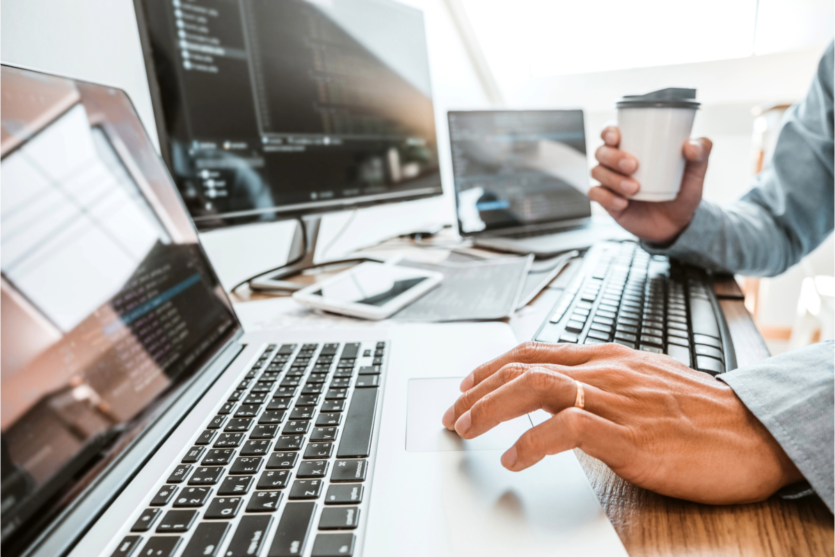 Person wearing a ring working on a laptop with code on screens, while holding a cup. Multiple computer monitors and devices are on the desk, showcasing Shopify Website Development to boost sales and conversions.