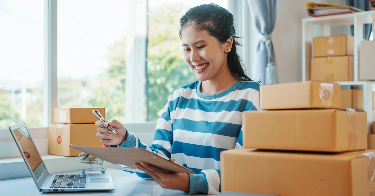 A woman smiles while working at a desk with several cardboard boxes, a laptop, and a clipboard, implementing efficient strategies for e-commerce returns.