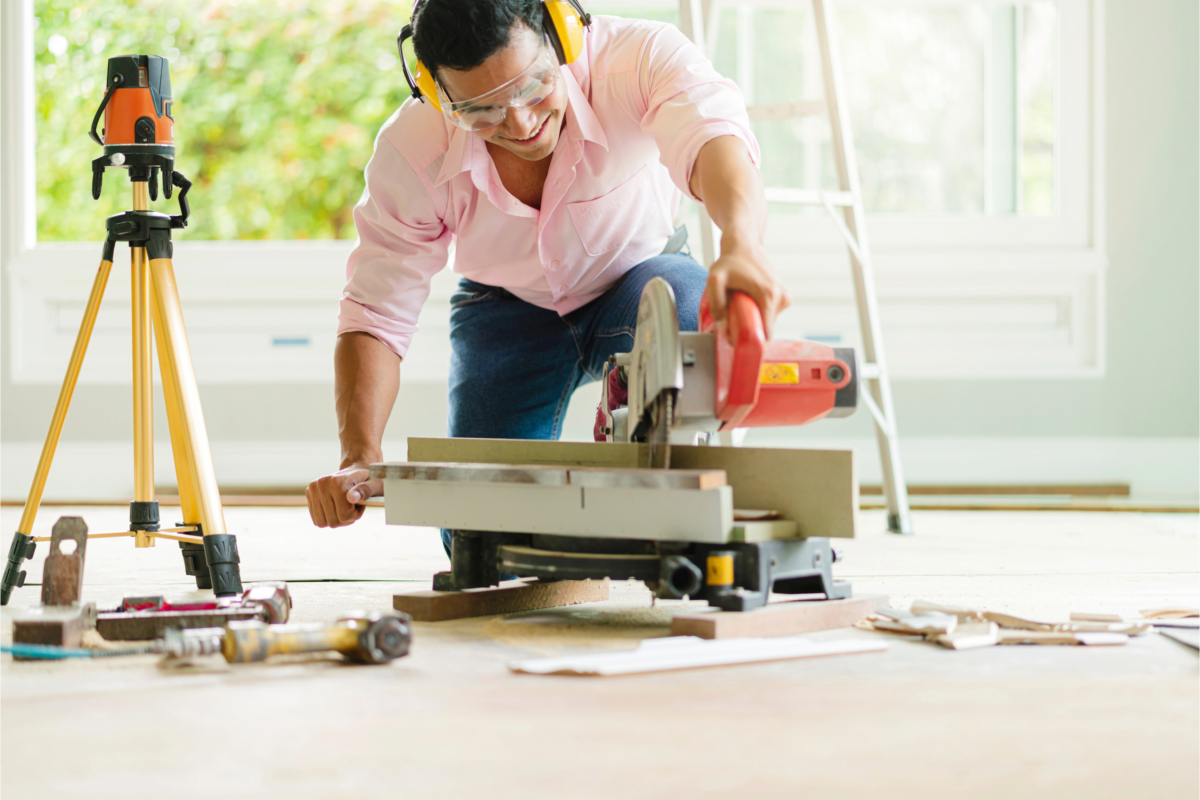 A person wearing safety gear operates a table saw to cut wood inside a room under renovation, with various tools and a ladder in the background, embodying the spirit of home improvement.