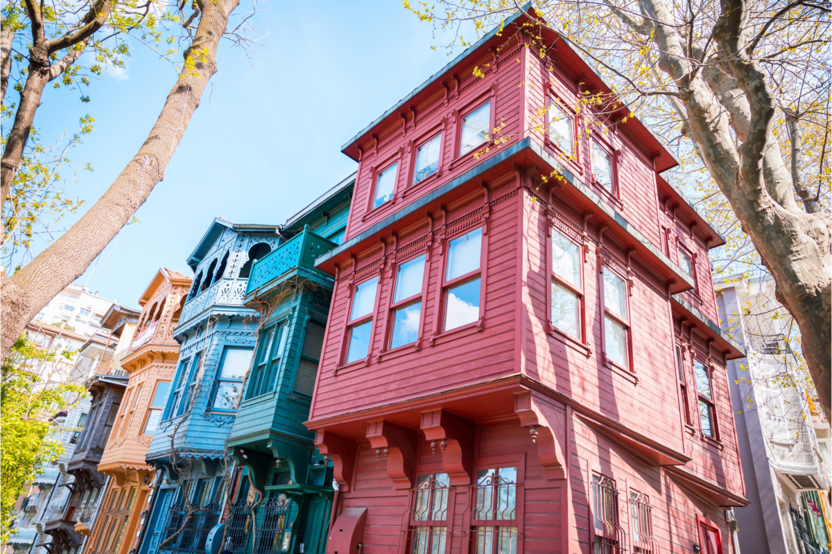 Row of colorful, historic wooden houses with large windows and ornate details under a clear blue sky, framed by trees in the foreground, offering attractive property investments with promising returns in Turkey.