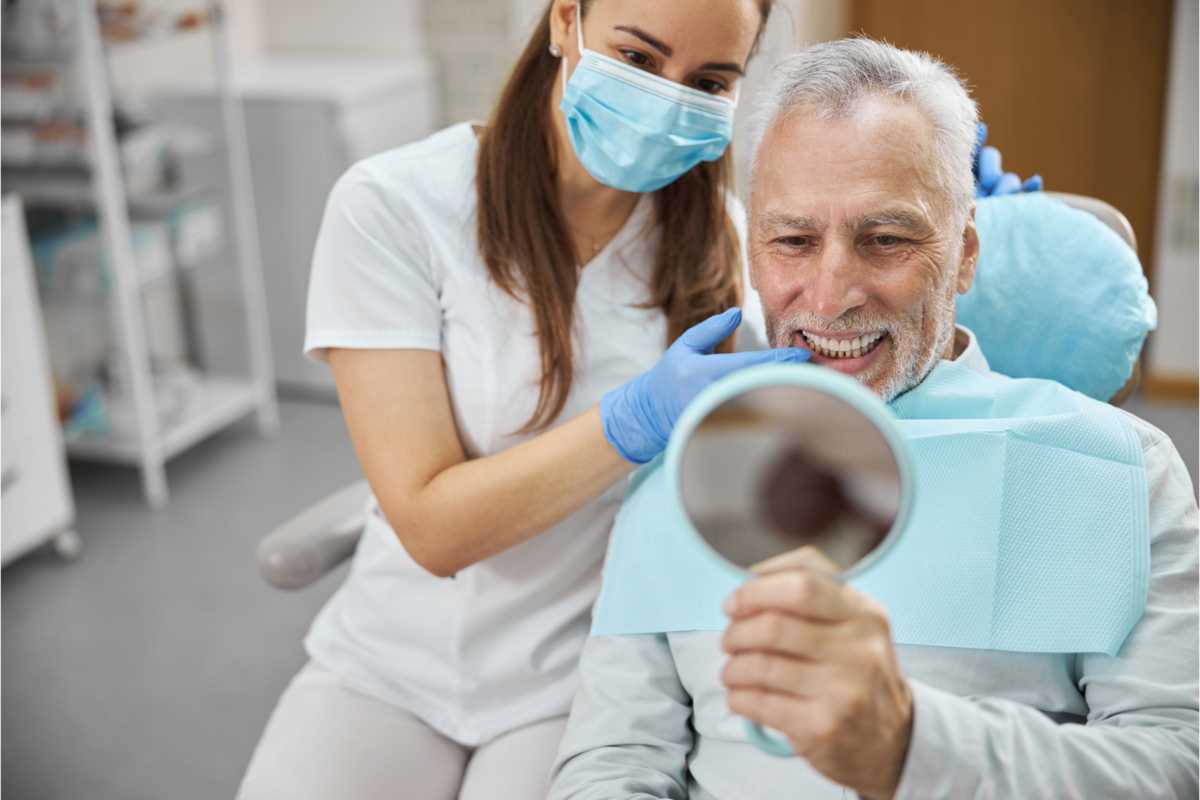 A dentist wearing a mask and gloves assists a smiling elderly man in a dental chair holding a mirror while examining his teeth, showcasing revolutionary advancements that mark a new era in dentistry.
