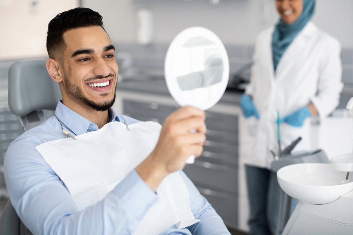 A man sitting in a dental chair smiles while admiring his perfect smile in a handheld mirror. An expert dentist stands in the background, smiling and wearing dental attire.