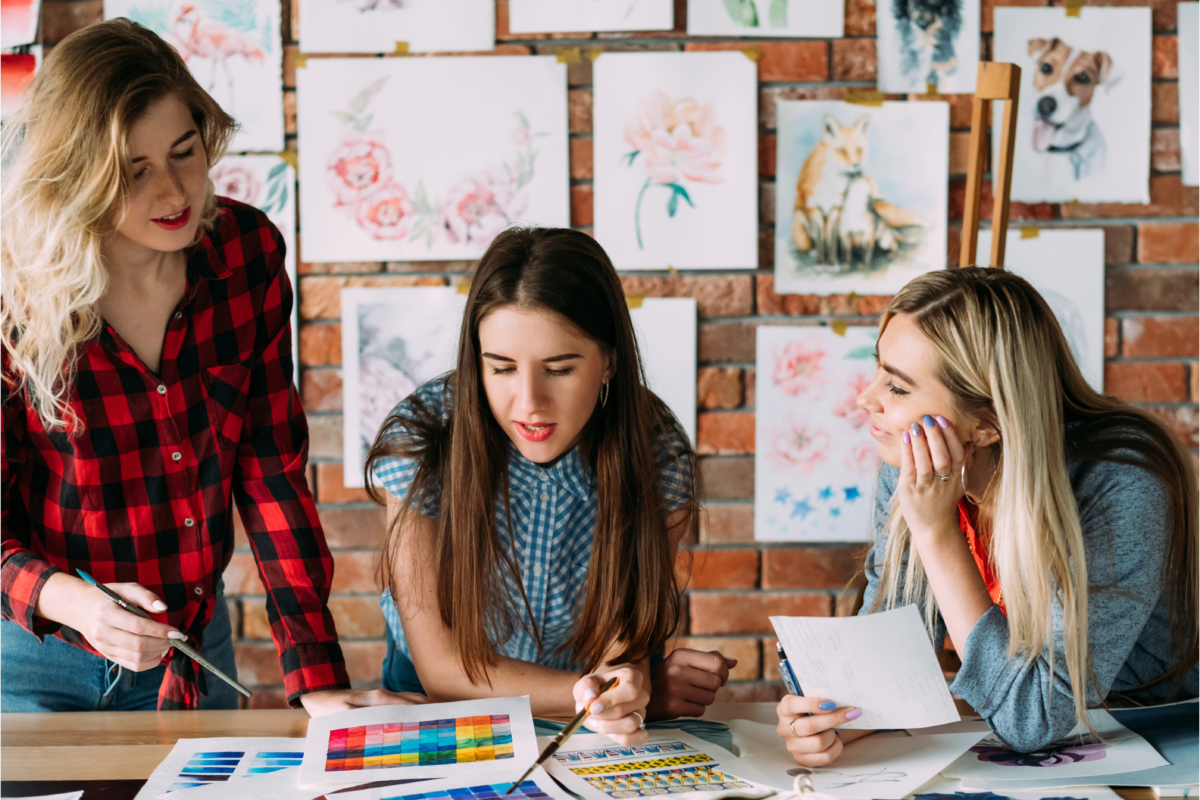 Three women in Melbourne collaborate on an art project at a table, unlocking their creative potential. They are surrounded by various watercolor paintings adorning the brick wall in the background, reminiscent of vibrant art courses.