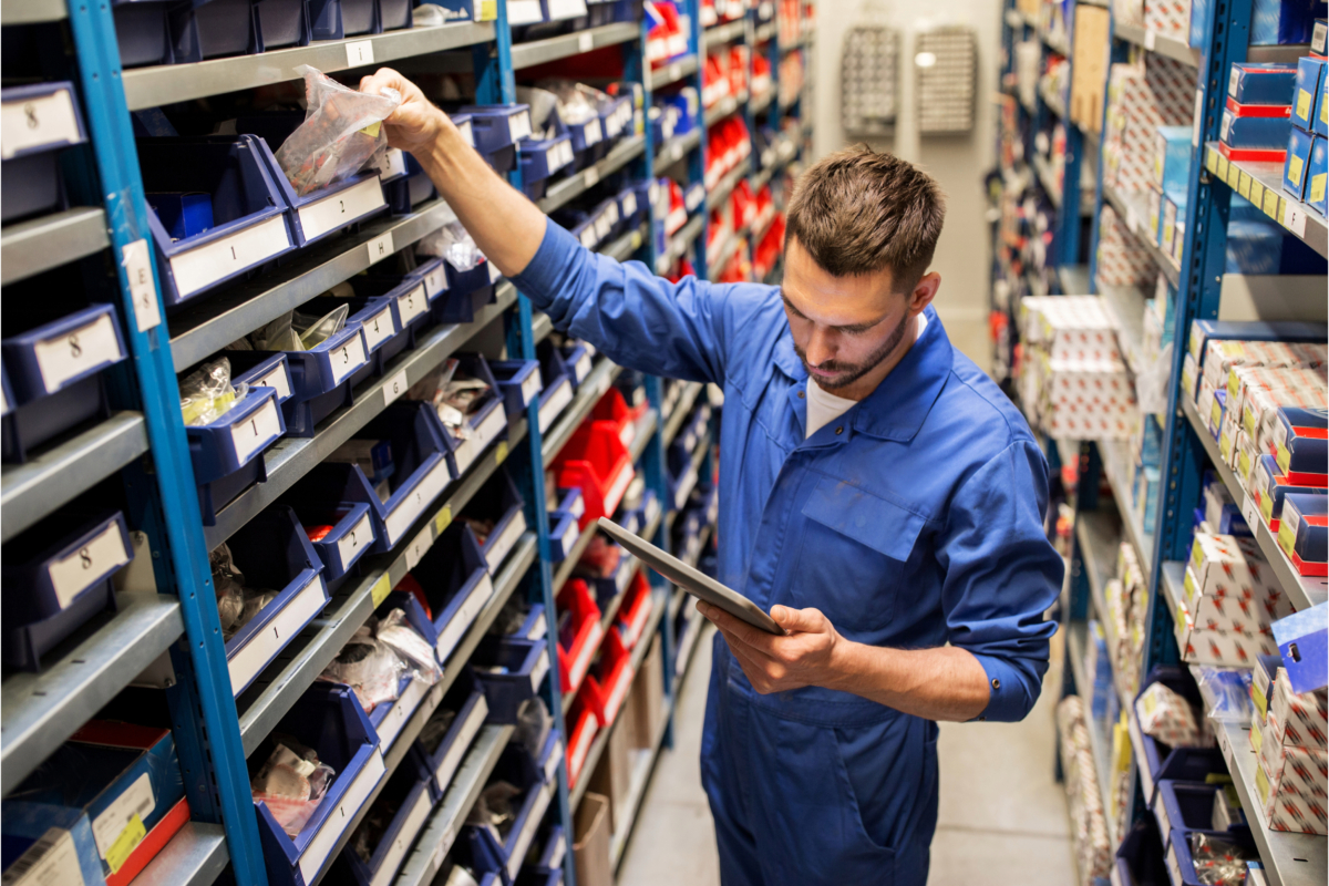 A man in a blue overall is standing in an aisle of shelves filled with small parts, using a tablet. He is picking out export automotive parts from a bin on one of the shelves.