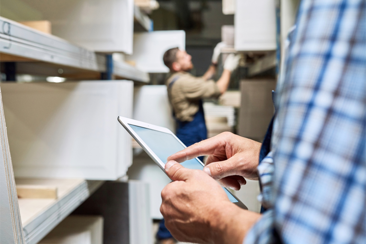 Person in the foreground uses a tablet, calculating shipping costs, while another person in the background organizes items on shelves with precision in a workspace.