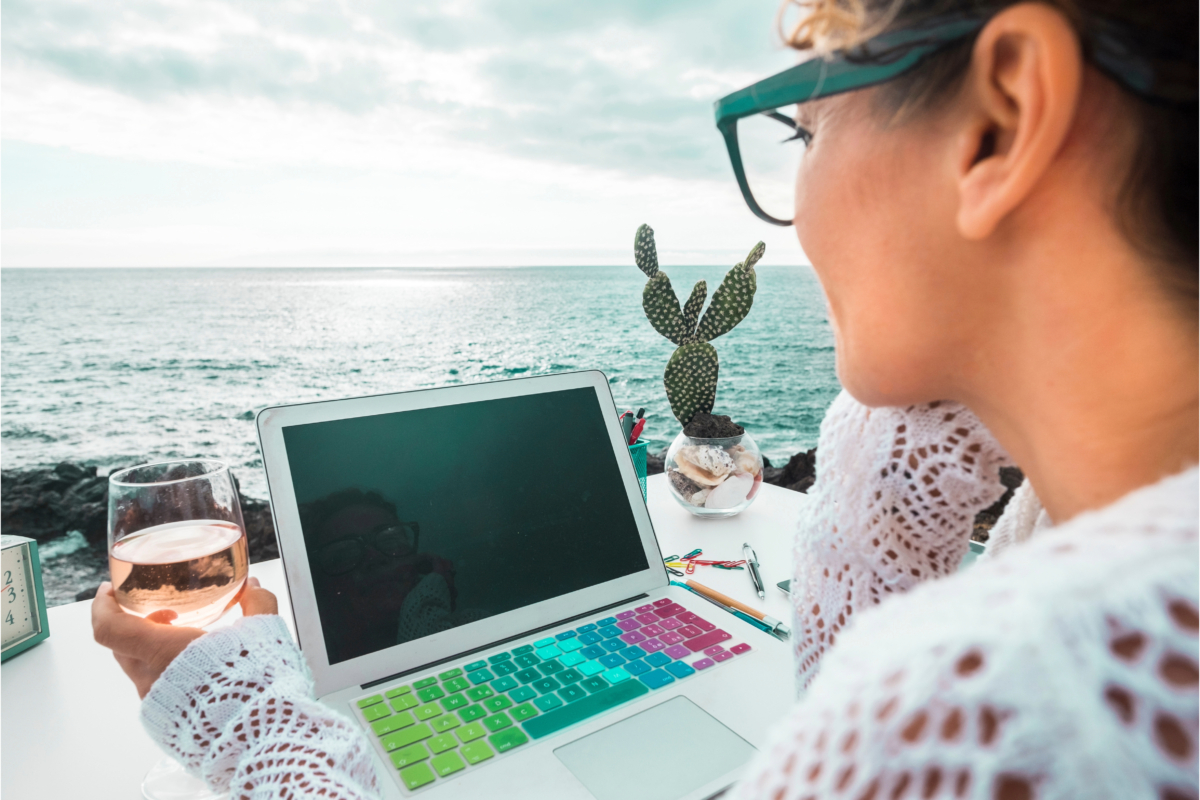 A female entrepreneur working remotely, while enjoying a glass of wine at a table.