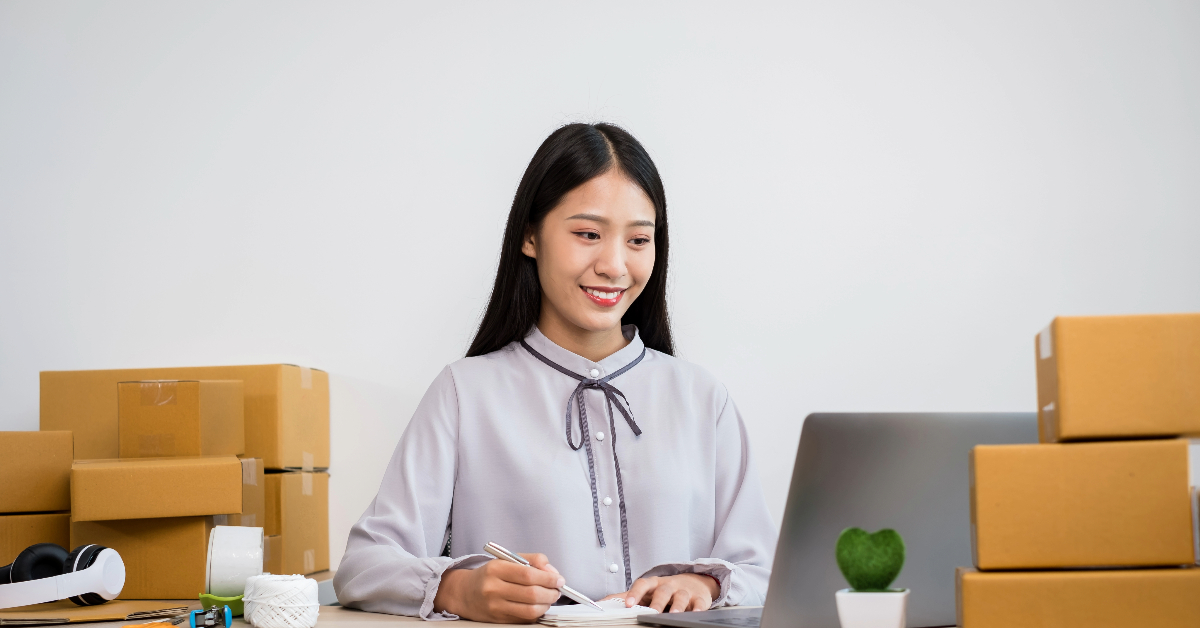 A woman sits at a desk with a laptop, pen, and notebook, surrounded by cardboard boxes—deep in thought about her ecommerce exit strategy.