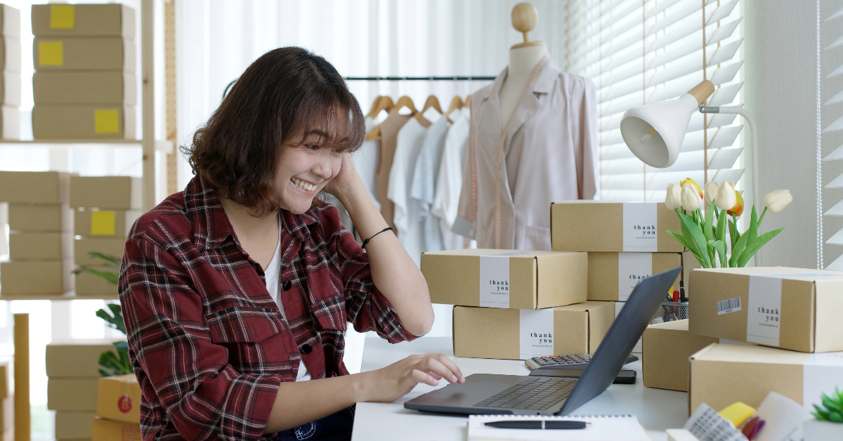 A woman is working on a laptop in her home office, surrounded by packing boxes and a clothing rack, efficiently managing her e-commerce business.
