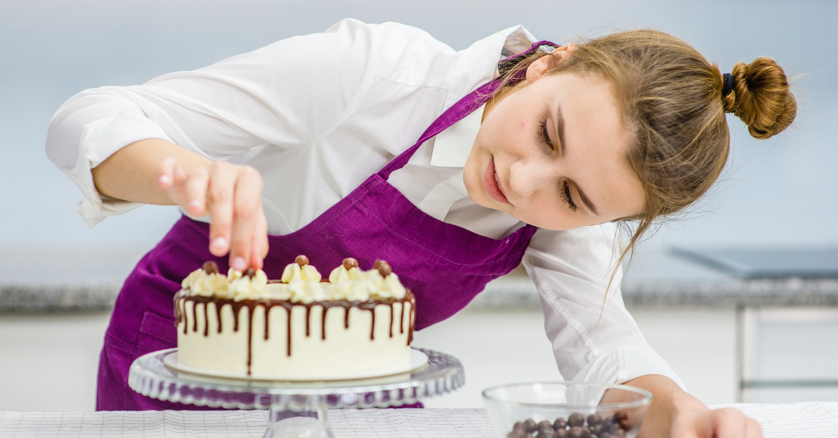 A girl in a purple apron enhancing a cake with chocolate, keeping up with the latest cake trends in the bakery business.