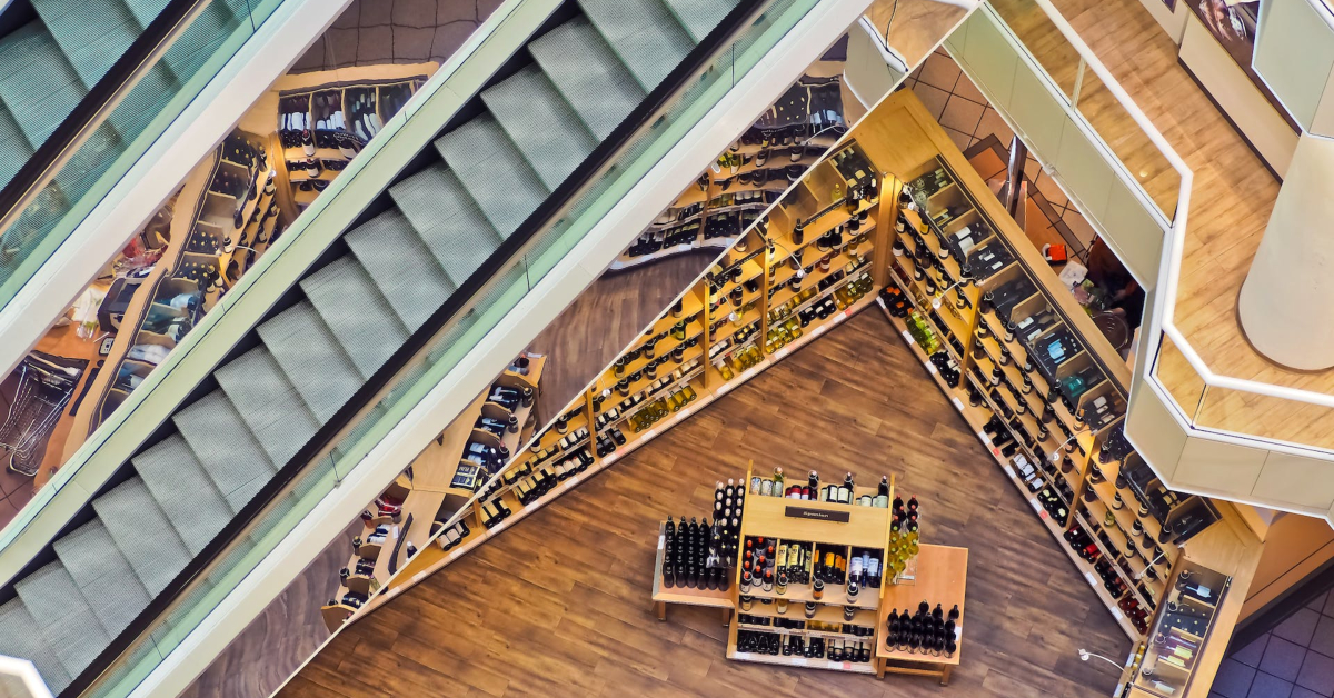 From a bird's-eye view, you can see the retail success of multiple escalators leading to a wine shop with bottles beautifully displayed on wooden shelves and a central counter. The light wood flooring adds to the store's inviting atmosphere.