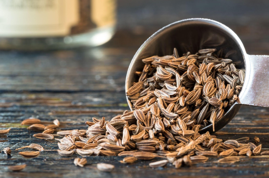 A metallic scoop is spilling spicy caraway seeds onto a rustic wooden surface, filling the air with their rich, aromatic smell.
