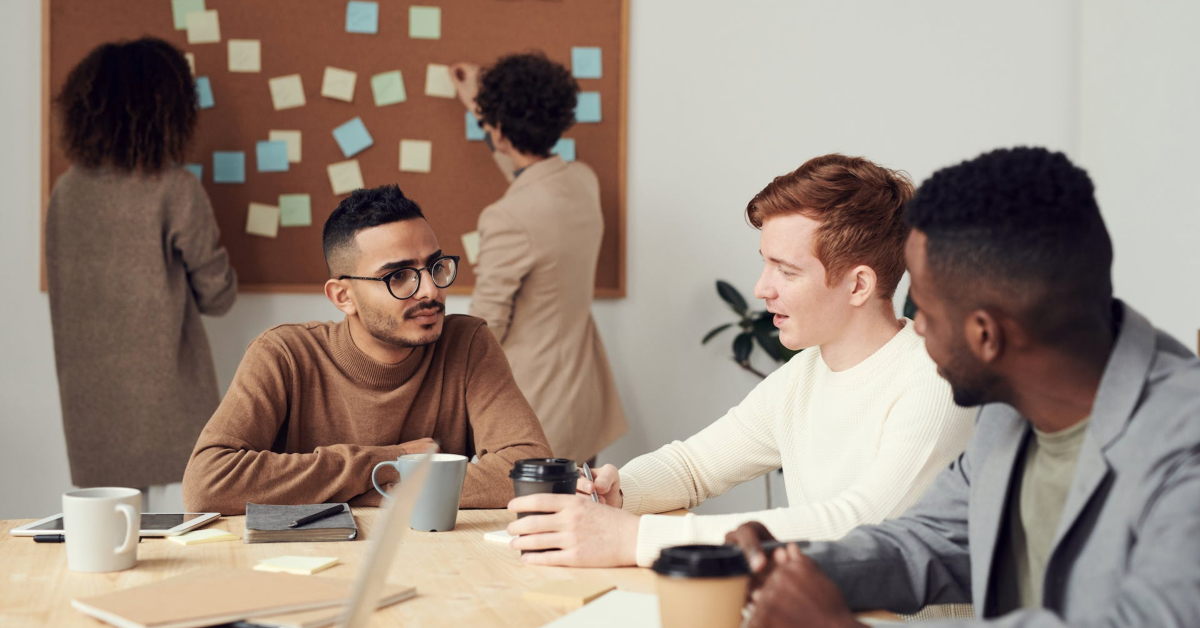 A group of people having a business discussion around a table with coffee cups, notebooks, and devices while others place sticky notes on a bulletin board in the background, showcasing effective communication.