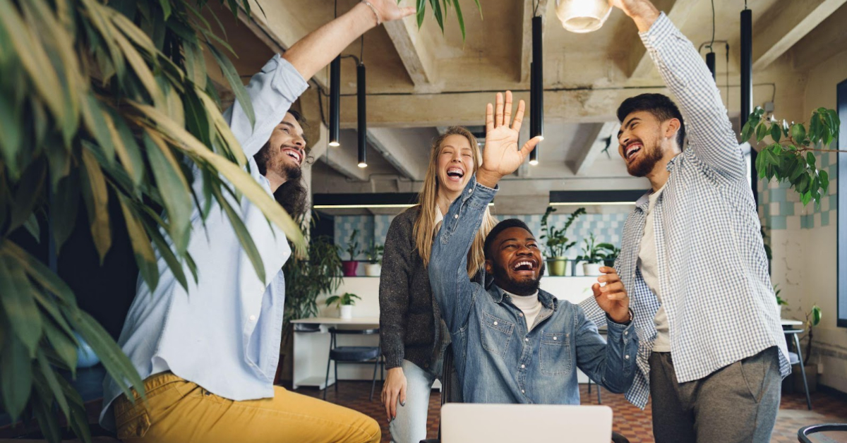 A group of four people stand together in an office, smiling and raising their hands in celebration. Plants and office furniture are visible in the background, creating a lively atmosphere that enhances the employee experience.