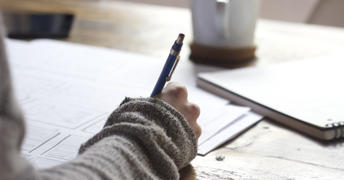 A person holding a pen, writing on papers placed on a wooden table. A notebook and a coffee mug are also visible on the table, as they prepare for their consulting case study interview.