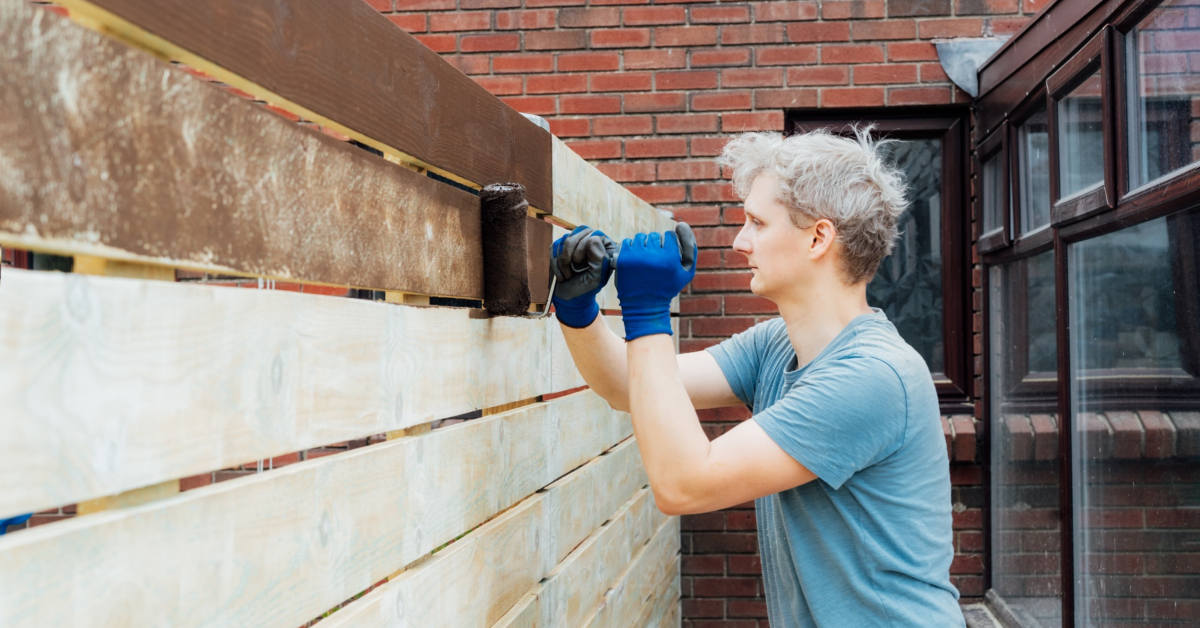 Person wearing a blue shirt and gloves, using a roller brush to paint a wooden fence brown outside a brick building as part of routine fence maintenance.