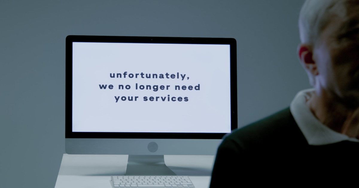 A man is standing in front of a computer screen that says he has unfortunately lost his job.