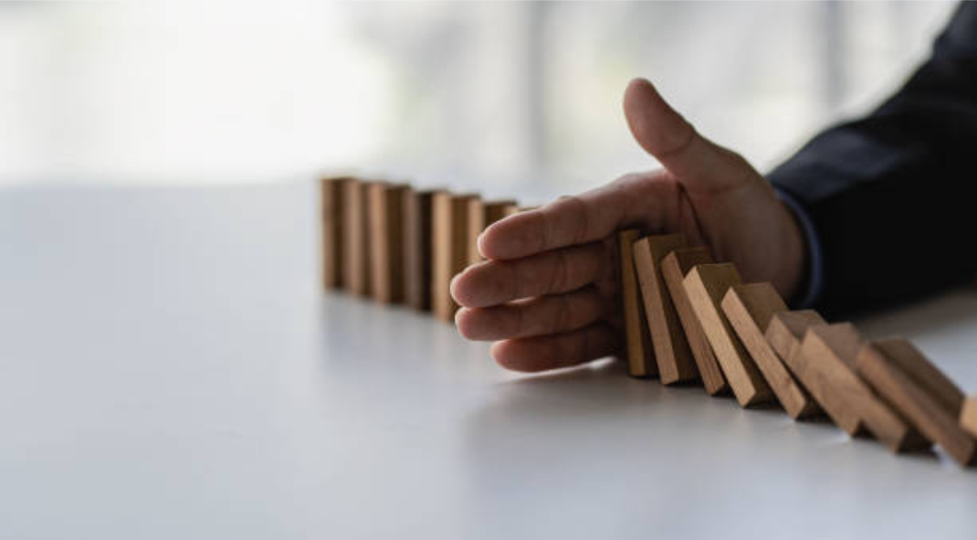 A hand stops a row of falling wooden dominoes on a white surface, demonstrating key skills in risk management to prevent them from continuing to topple over.