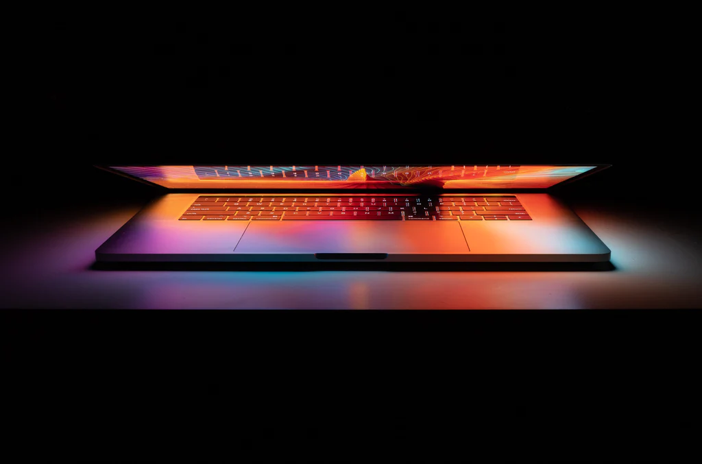 A laptop with colorful lights on it is shown in a dark room, highlighting the vibrant display and illuminating the surrounding atmosphere.