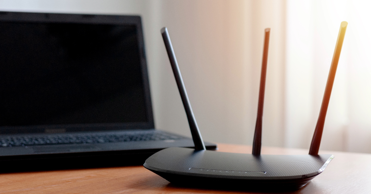 A black laptop is placed on a wooden desk next to a black wireless router with three antennas, ready to deliver high-speed connectivity from Metronet, the best internet provider in Tallahassee.