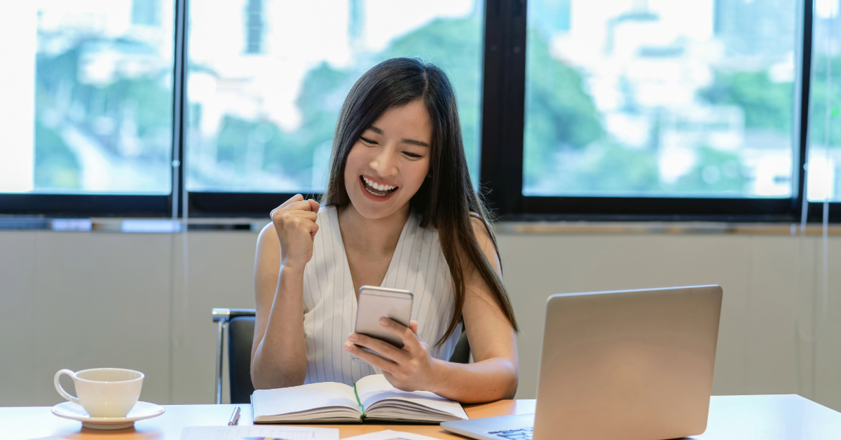 A woman sits at a desk with a laptop, notebook, and a cup of coffee. Smiling and raising her fist while looking at her smartphone, she has just discovered one of the best investment apps for beginners.