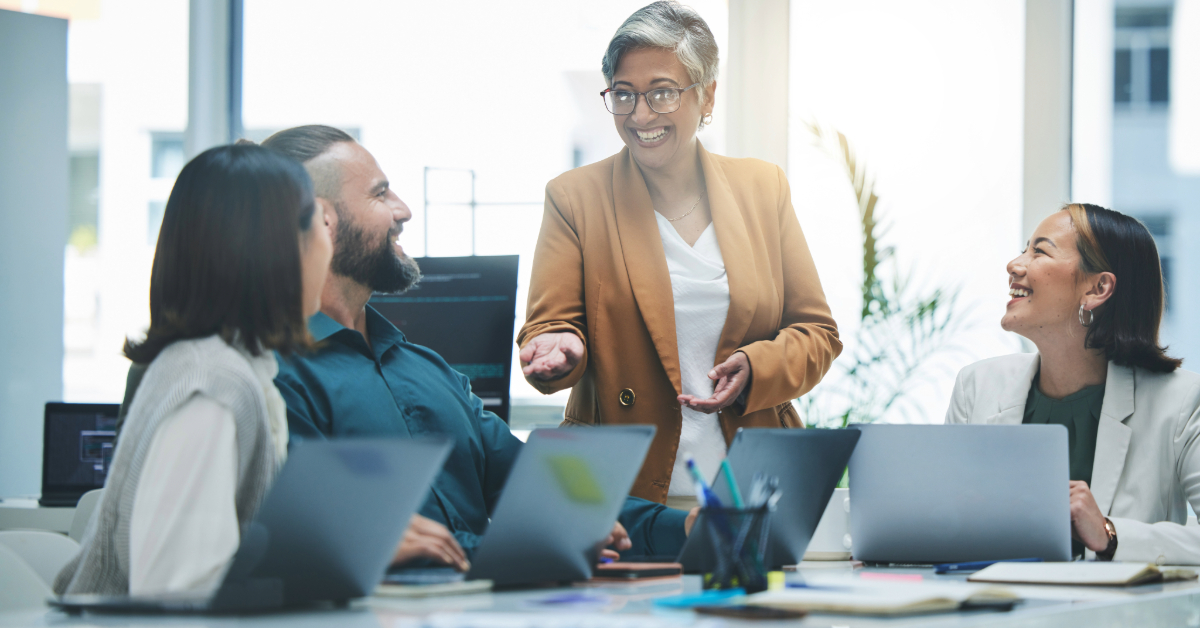 A group of four people in a meeting room, seated with laptops open. One person is standing and speaking, offering workflow tips, while the others are smiling and listening attentively. The atmosphere reflects their dedication to improving company productivity.