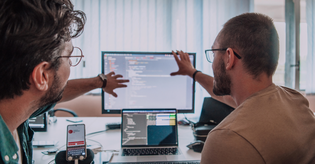 Two men review code on a large monitor in an office setting, with laptops and a smartphone on the table in front of them, showcasing their consulting expertise in software development services.