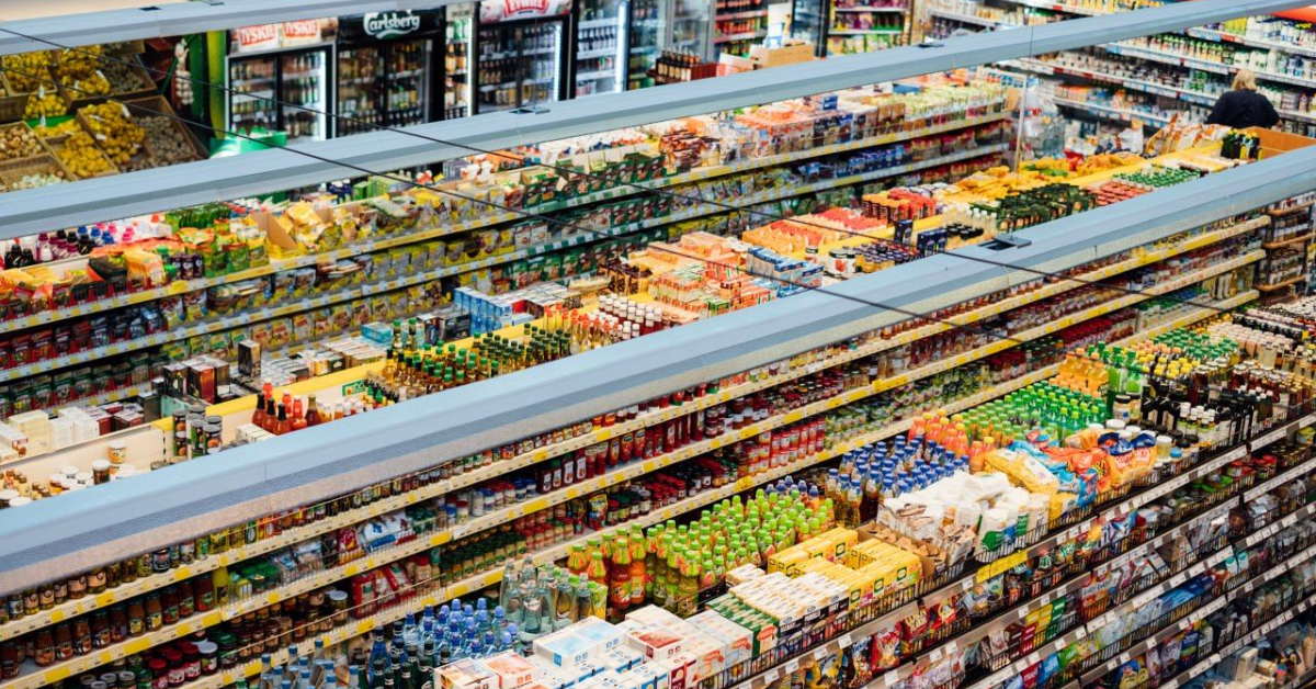 A large supermarket aisle filled with various products on shelves, including canned goods, snacks, beverages, and other food items. The shelves are fully stocked and organized, reflecting the 2024 trends in the future of shopping.