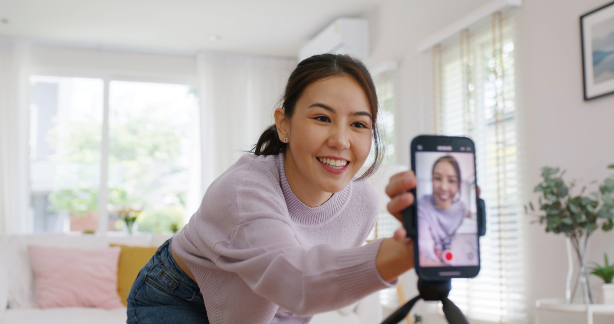 A woman in a purple sweater smiles as she adjusts her phone on a tripod, preparing to record or stream inside a well-lit room with large windows and plants, perhaps ready to share principles of viral marketing.