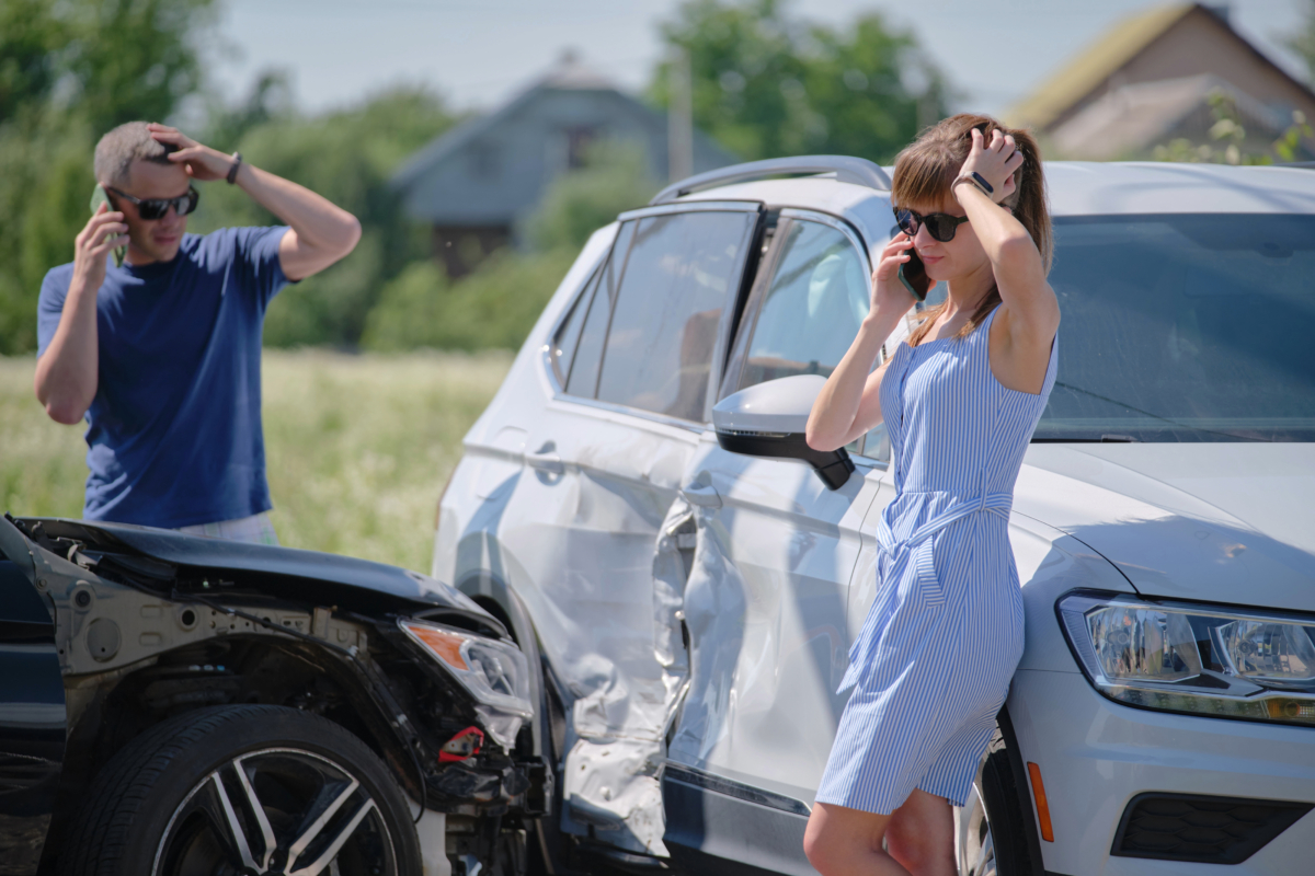 Two people on phones stand beside their damaged vehicles after a car accident, appearing distressed. The white SUV shows significant side damage while the black car's front end is damaged. This scene highlights the potential need for personal injury settlements following such incidents.