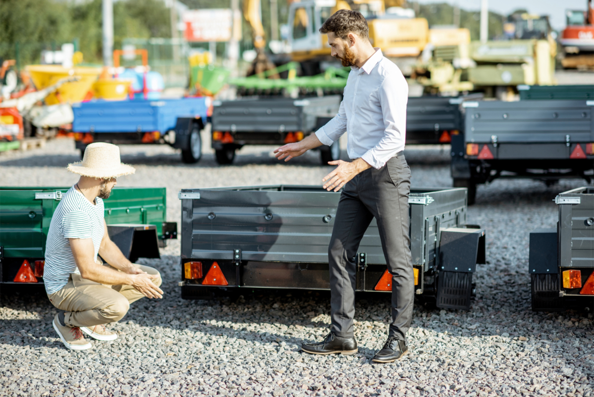Two men examine trailers parked in an outdoor lot; one is crouching and the other standing, gesturing towards a trailer. Machinery and equipment are visible in the background. They seem to be reviewing a trailer buying checklist while considering the used trailers for sale.