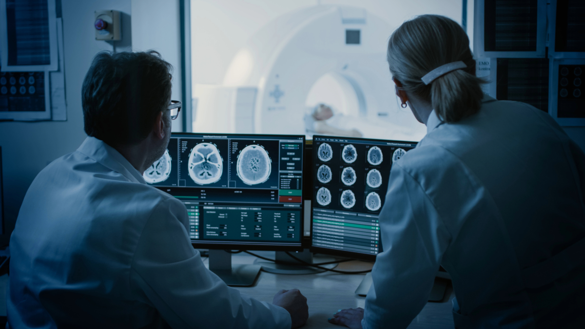 Two medical professionals in lab coats examine brain MRI scans on monitors, utilizing advanced AI radiology software, with a patient undergoing an MRI scan in the background.