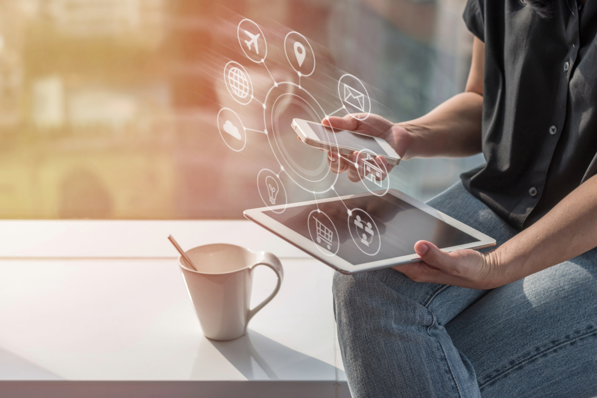 A person, exuding creativity, sits while using a smartphone and tablet, surrounded by various digital icons like an airplane, shopping cart, and email floating around the devices. A cup with a spoon rests on the table nearby.