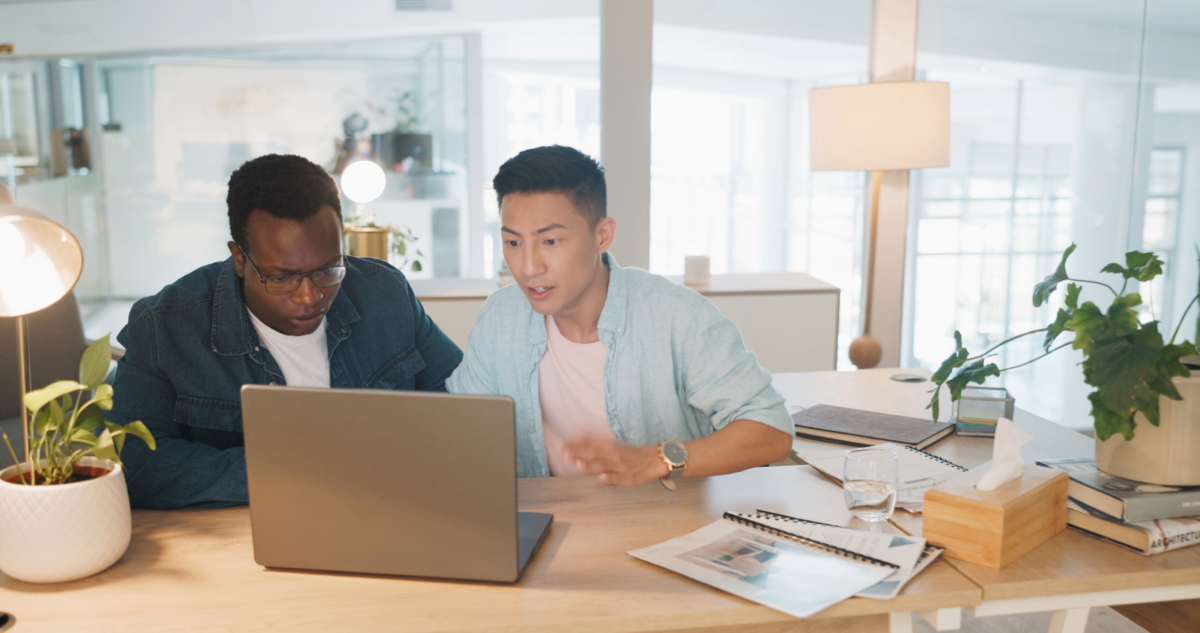 Two people sit at a desk working on a laptop in a bright office, with notebooks, documents, and plants nearby. They focus on WordPress safety while discussing how to safeguard your site from security risks.