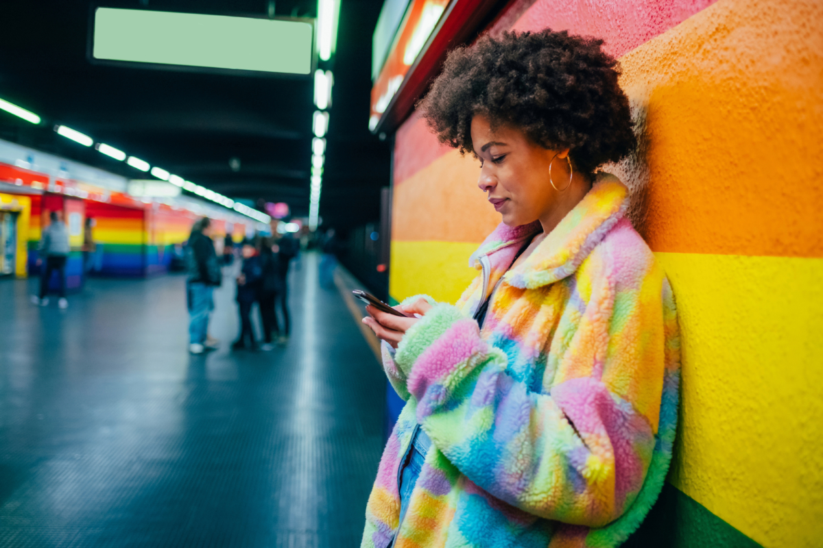 A person in a colorful rainbow fur coat leans against a rainbow-striped wall in a subway station, looking at their phone. Transforming the scene with vibrant hues, they navigate their online presence, while other people move in the background.