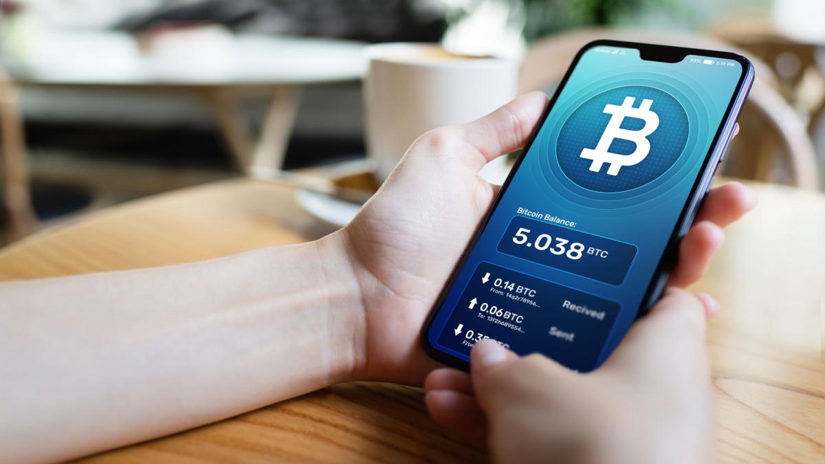 Person holding a smartphone displaying a cryptocurrency wallet with a balance of 5.038 BTC, illustrating the growing use of cryptocurrency payments across various industries. A cup and blurred background are visible in the background.