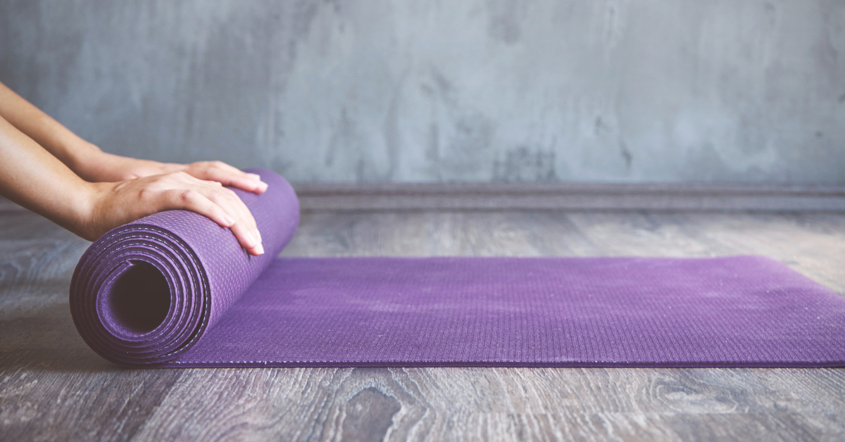 Hands rolling up a personalized purple yoga mat on a wooden floor against a gray textured wall, showcasing the power of personalized yoga mats.