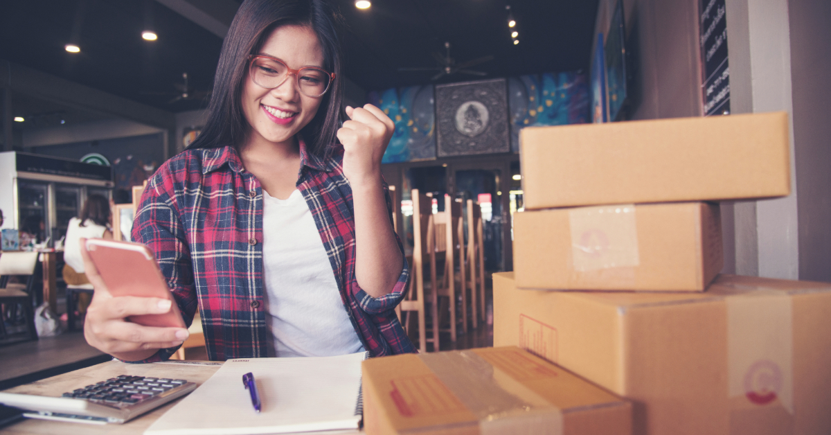 Una donna è seduta a un tavolo con delle scatole davanti a sé e sta esaminando attentamente le informazioni sui prezzi dei prodotti per i venditori di Amazon.