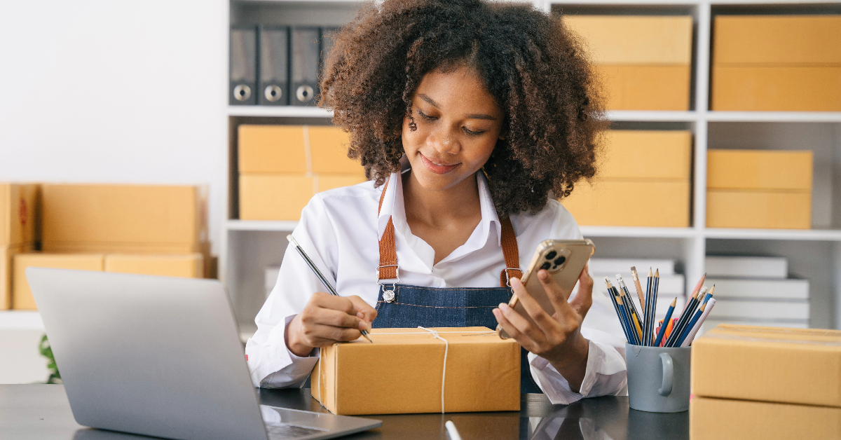 A woman working at a desk with boxes and a cell phone, managing the shipping of books internationally.